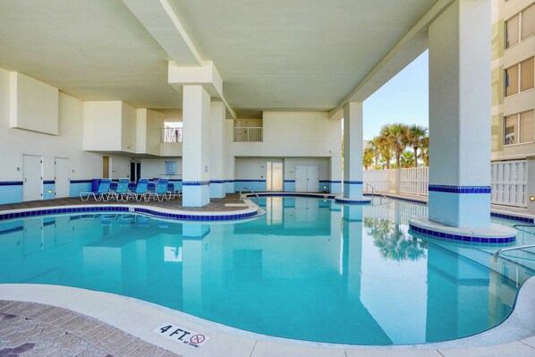 A large indoor swimming pool with clear blue water, surrounded by white pillars and blue-tiled edges. There are lounge chairs along the poolside and a small upper deck area.