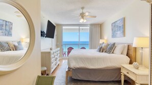 Une chambre d’hôtel avec un grand lit, un ventilateur de plafond, un téléviseur fixé au mur, un balcon avec vue sur l’océan et une chaise près de la fenêtre.