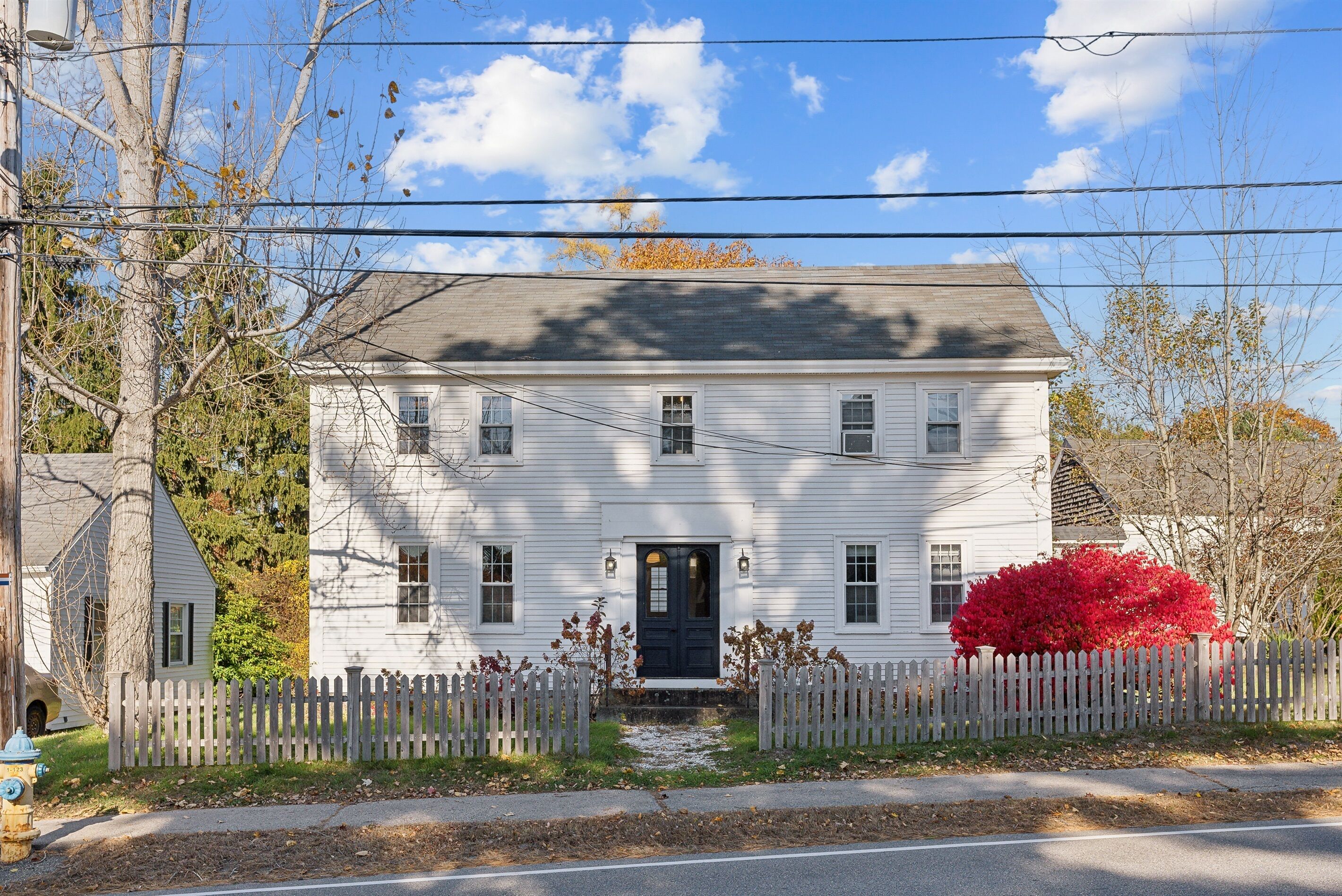 A white two-story house with a black front door, a picket fence, and a red bush in front.