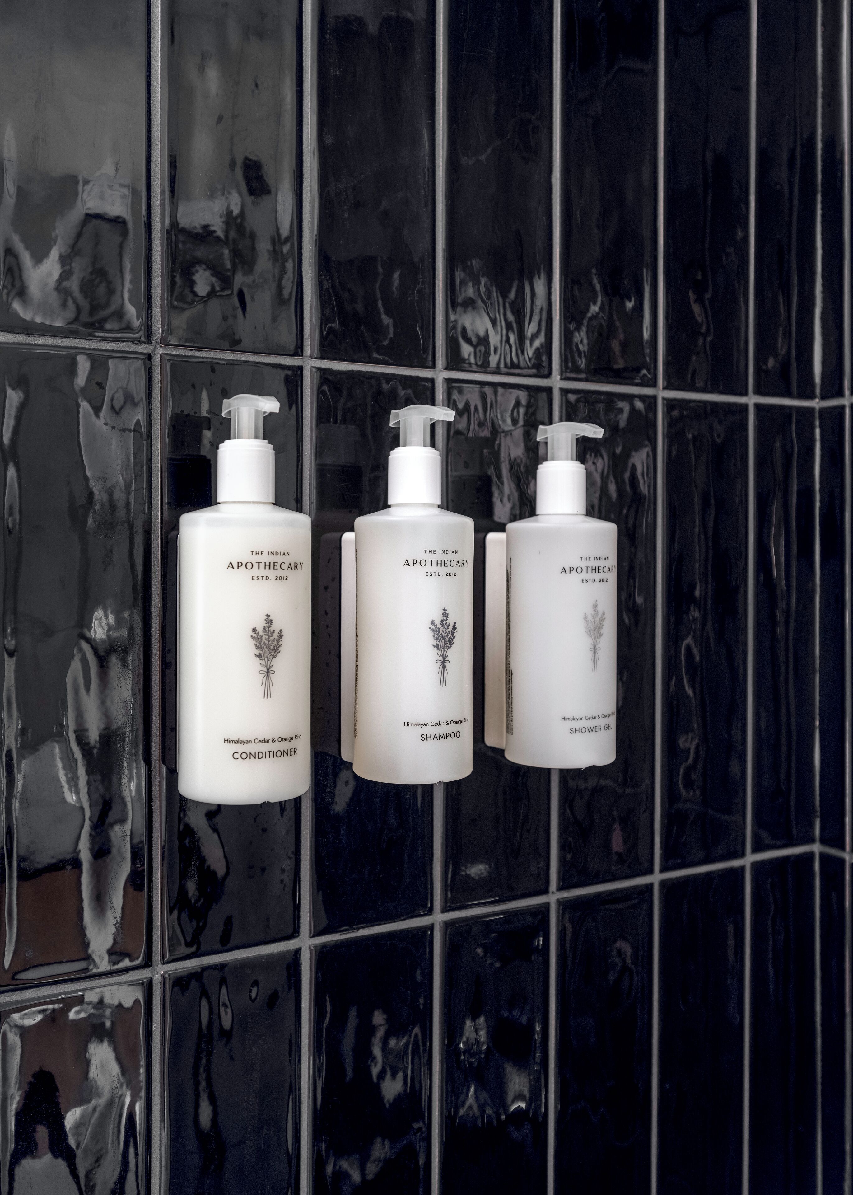 Three bottles of The Indian Apothecary Himalayan Cedar & Orange Oil Shower Gel on a tiled wall.