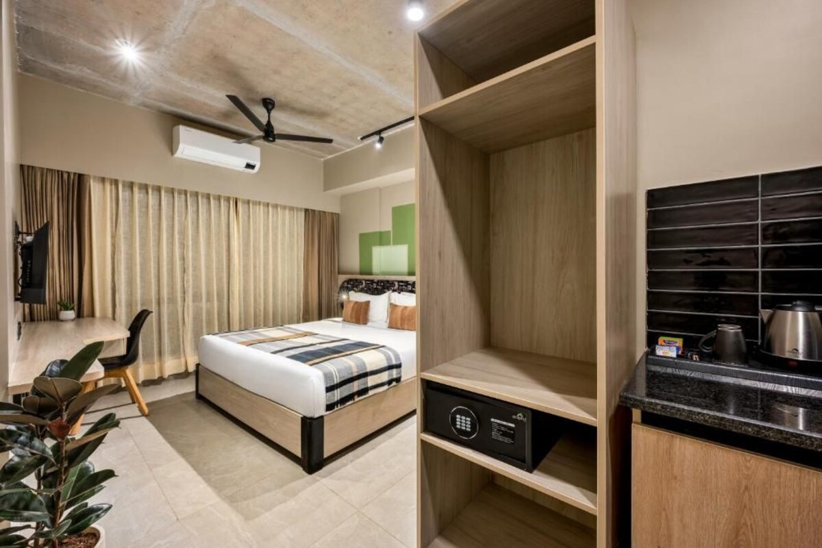 Een hotelkamer met een bed, een bureau, een stoel, een plant, een plafondventilator en een kitchenette.