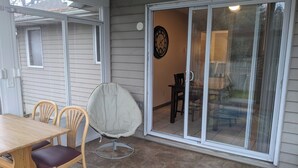Un espacio de patio cubierto con mesa y sillas de madera, una silla acolchada y un ventanal corredizo que da a un ambiente interior.
