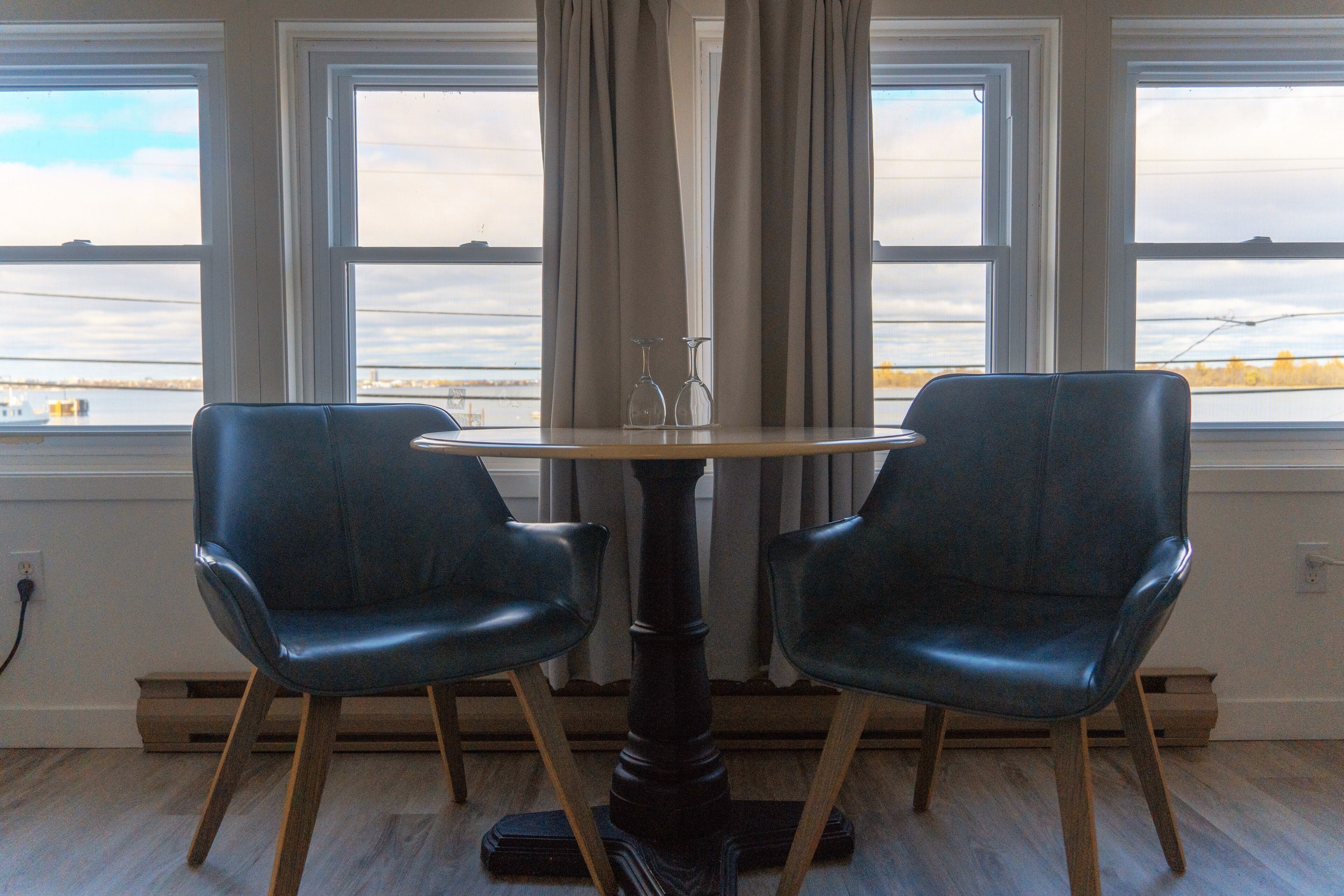 A room with a round table and two chairs, large windows with a view of a body of water, and a curtain partially drawn.