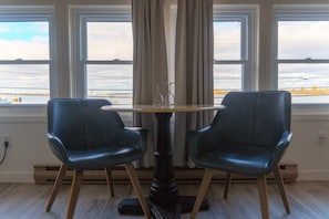 Ein Zimmer mit einem runden Tisch und zwei Stühlen, großen Fenstern mit Blick auf ein Gewässer und einem teilweise geöffneten Vorhang.