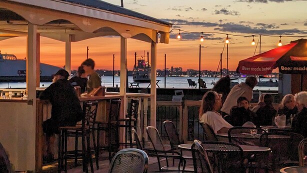 An outdoor seating area with tables and chairs, overlooking a body of water with a city skyline in the background. - Hotel Wolfe Island (Wolfe Island)