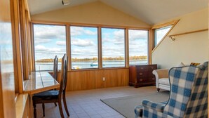 A room with a large window overlooking a lake, a dining table with chairs, a sofa, and a wooden cabinet. - Hotel Wolfe Island (Wolfe Island)