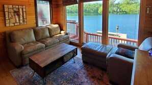 Un acogedor interior de cabaña con techo de madera, un sofá, una mesa de centro y vista a un lago a través de amplios ventanales.