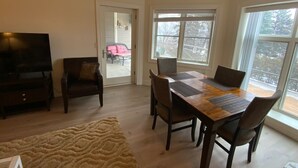 A living room with a TV, a dining table with chairs, a sofa, and a large window with a view of trees. - Lovely 2-bedroom Suite with lots of Amenities (Traverse City)