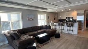 A modern living room with a sectional sofa, a coffee table, and a rug. There is a kitchen area with bar stools and a refrigerator.