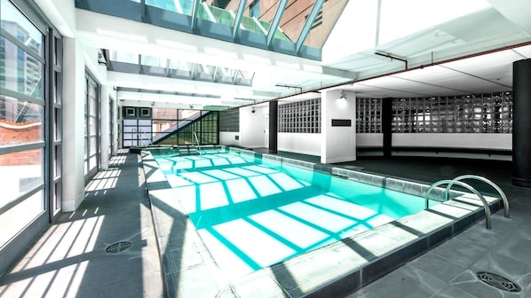 A modern indoor swimming pool with clear glass walls, a sunlit interior, and a view of the outside.