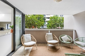 A balcony with wicker chairs, a small table, and a view of trees and buildings.