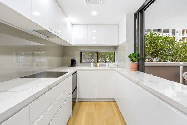 A modern kitchen with white cabinetry, a black induction cooktop, and a window with a view of greenery outside.