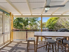 Eine Terrassengruppe im Freien mit einem Holztisch und Stühlen, einem Deckenventilator und Blick auf Bäume sowie ein Gebäude im Hintergrund.