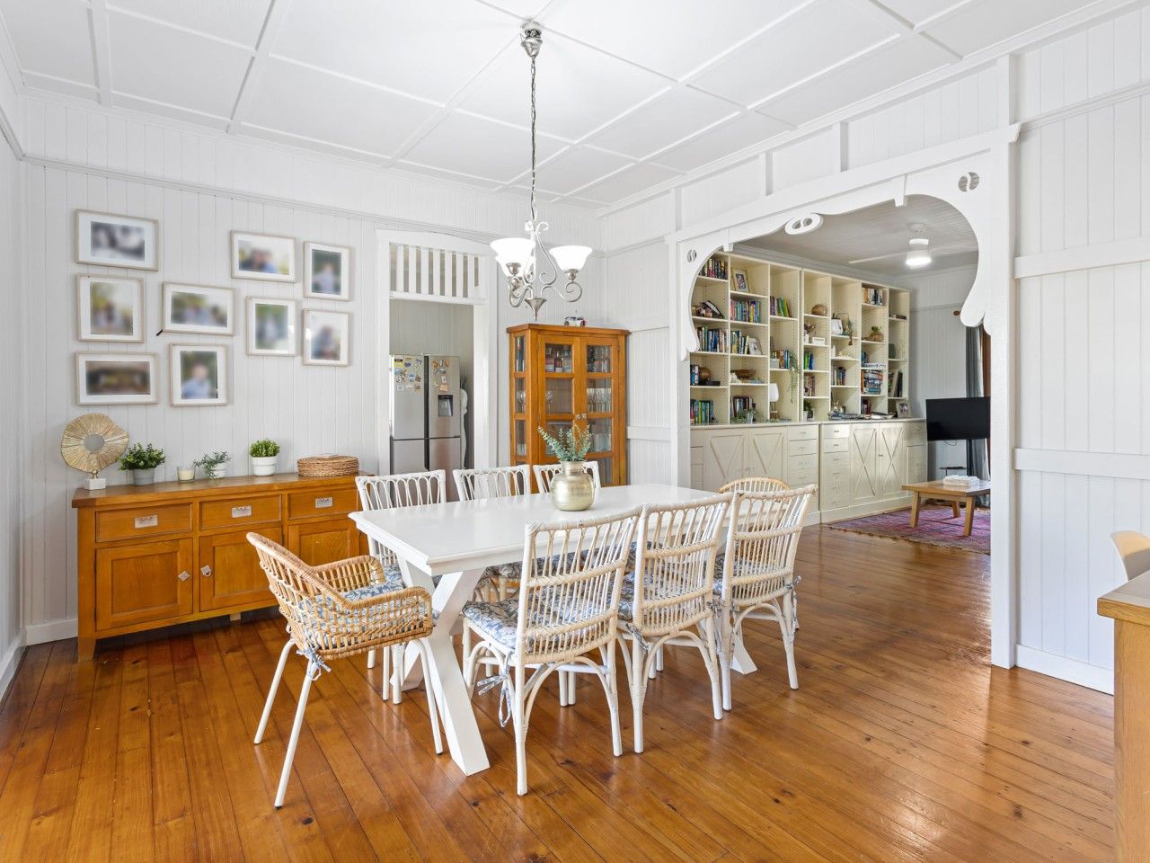 A dining area with a white table and wicker chairs, a wooden cabinet, and a bookshelf in the background.