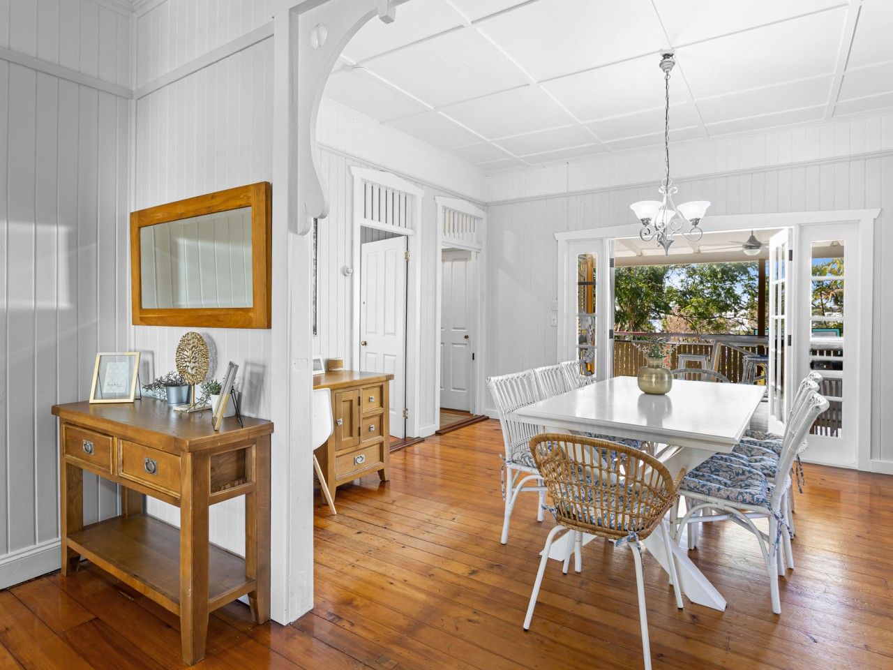 A dining area with a white table and wicker chairs, a wooden sideboard, and a mirror on the wall.