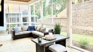Un patio cubierto con muebles de mimbre, una mesa con adornos y ventanas amplias que ofrecen vistas del jardín trasero y los alrededores.