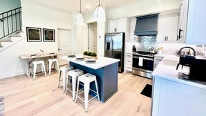 Una cocina moderna con una isla central, armarios blancos, electrodomésticos de acero inoxidable y una escalera que sube a un nivel superior.