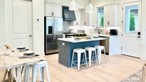 Una cocina moderna con armarios blancos, una isla central con taburetes, electrodomésticos de acero inoxidable e iluminación colgante.