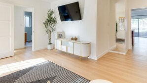 Ein modernes Wohnzimmer mit einem Wand-Fernseher, einer Topfpflanze, einem Konsolentisch mit Dekoration und einem großen Fenster, das für natürliches Licht sorgt.