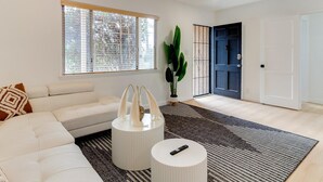 Ein modernes Wohnzimmer mit einem weißen Sofa, einem runden Couchtisch und einem großen Fenster mit Jalousien.