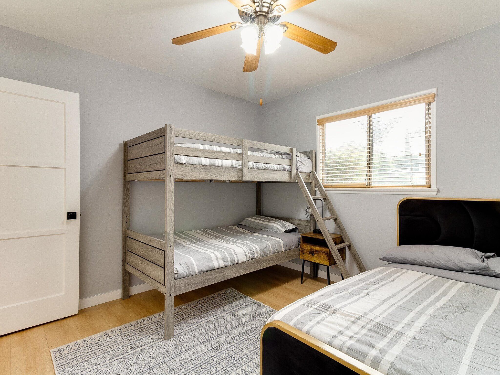 A bedroom with a bunk bed, a single bed, a ceiling fan, a window with blinds, and a patterned rug.