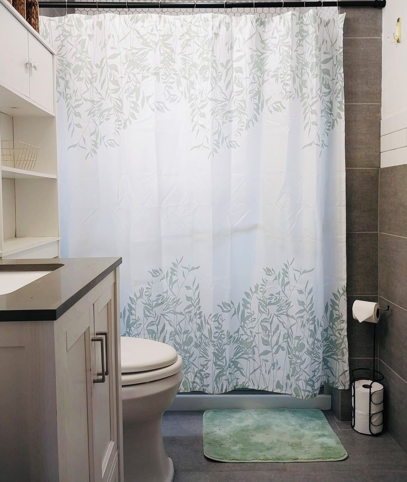 A bathroom with a toilet, a green rug, a shower curtain with a leaf pattern, and a towel rack.