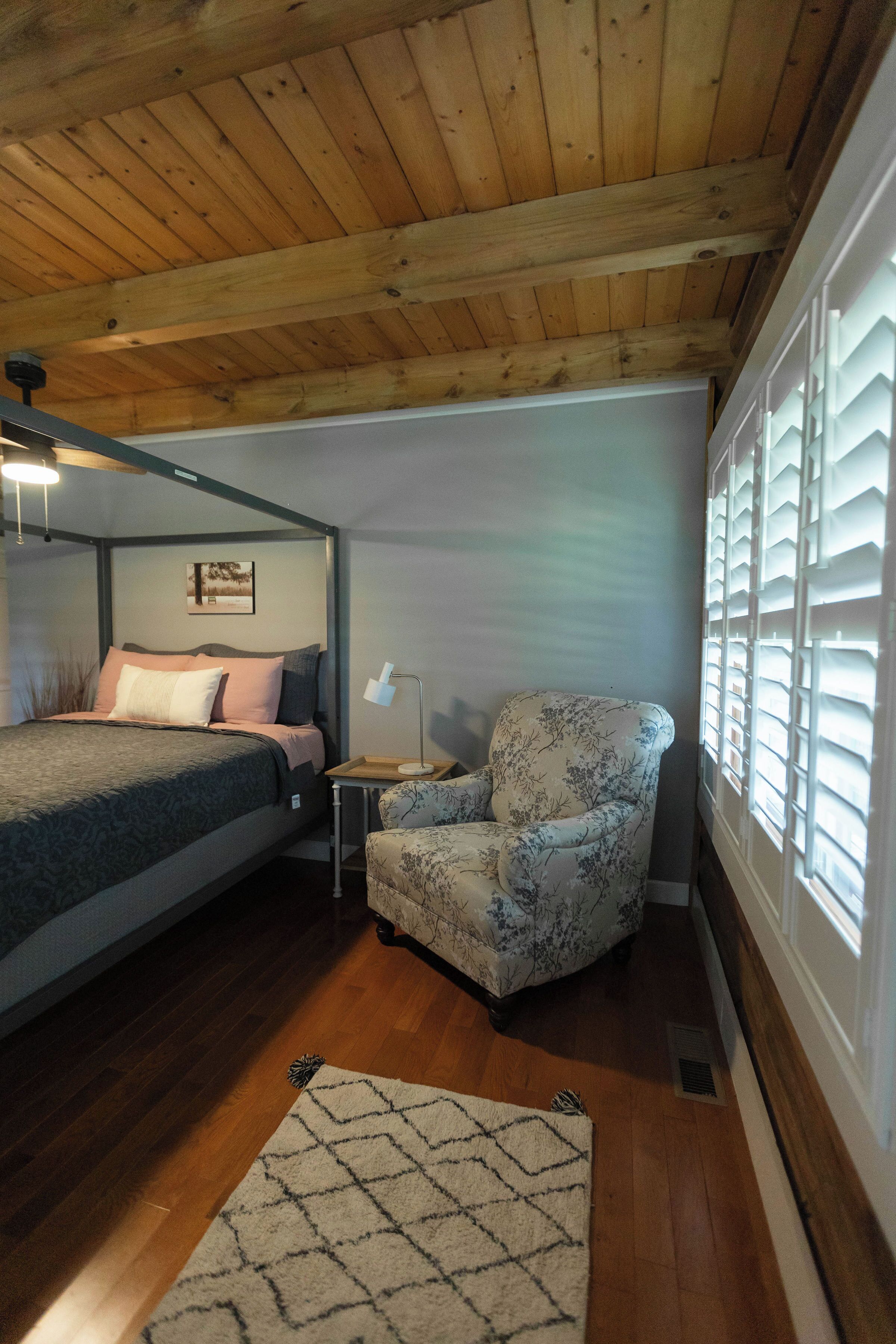 A bedroom with a wooden ceiling, a bed with a patterned bedspread, a chair with a floral design, a bedside table with a lamp, and a window with shutters.