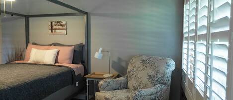 A bedroom with a wooden ceiling, a bed with a patterned bedspread, a chair with a floral design, a bedside table with a lamp, and a window with shutters.
