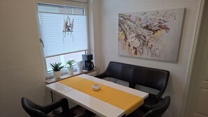 A dining area with a white table, two black chairs, a yellow placemat, a white cup, a coffee maker, and a plant on the windowsill. - Vacation apartment "Schöne Aussicht" with large balcony-terrace (Nordenham)
