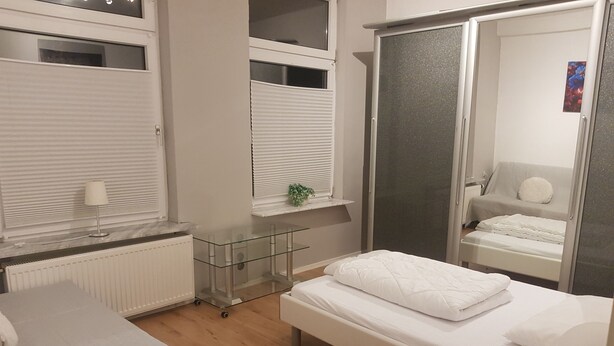 A bedroom with a bed, a bedside table, a window with blinds, a radiator, and a small shelf with a plant. - Vacation apartment "Schöne Aussicht" with large balcony-terrace (Nordenham)