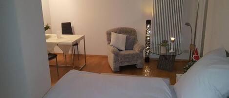 Una habitación de hotel con cama, un sillón pequeño, una mesa con lámpara y una ventana con persianas.