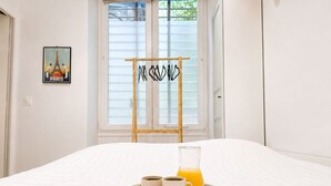 Una habitación de hotel con cama, ventana con persianas, escalera de madera, bandeja con dos tazas y un vaso de jugo de naranja, y un póster de la Torre Eiffel.