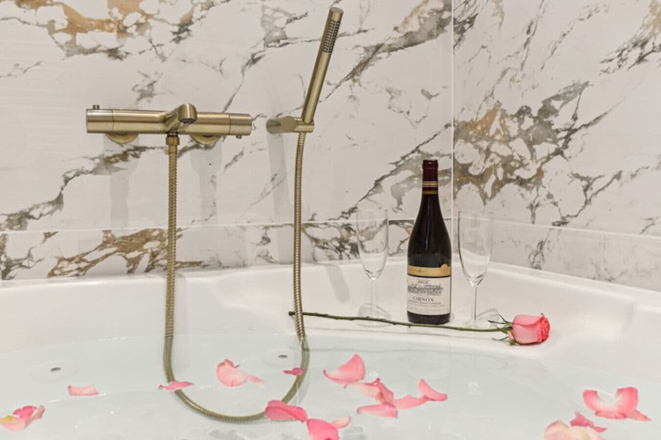 A bathtub with a showerhead and a bottle of wine, surrounded by rose petals.