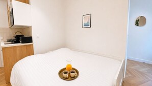 A neatly made bed with a tray of orange juice and two cups, a small framed picture on the wall, and a kitchen area with a coffee maker.