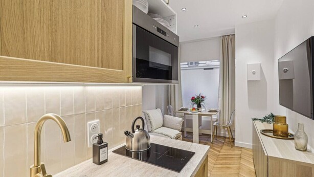 A modern kitchen with a sink, kettle, and stove, a dining area with a table and chairs, and a TV mounted on the wall.