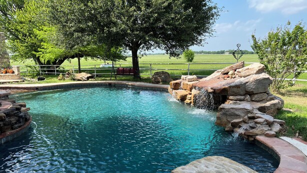 Una piscina con una cascada, rodeada por una terraza de piedra y un árbol.