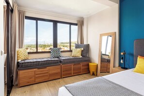 A bedroom with a large window, a bed, a bedside table, and a mirror.