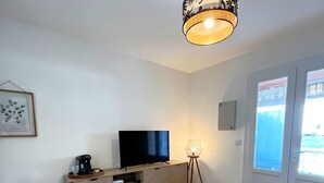 Een moderne woonkamer met een flatscreen televisie op een houten kast, een klein rond tafeltje met een kopje, een vloerlamp en een plafondlamp.