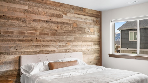 Un dormitorio con un panel de pared de madera, una cama con ropa de cama blanca y una ventana con vistas a una casa y montañas.