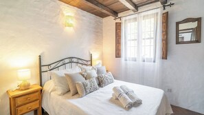 Een slaapkamer met een bed, kussens, een nachtkastje, een lamp, een raam met gordijnen en een spiegel aan de muur.