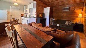 A cozy cabin interior with a wooden dining table, leather sofa, and a kitchen area in the background. - Mara Spring Lodges - Lodge 1 (Mara)