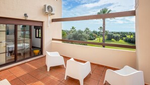 A balcony with three white chairs, a glass door leading to a room, and a view of a well-maintained garden.