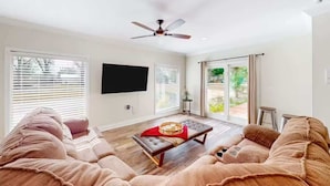 A spacious living room with a large flat-screen TV, a ceiling fan, and a sliding glass door leading to a patio. - Pool, Beaches, OWA, Dining, and More | Palmaway (Foley)