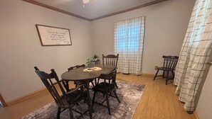 Une salle à manger avec une table ronde, six chaises, un tapis, une plante en pot et une fenêtre avec des rideaux.
