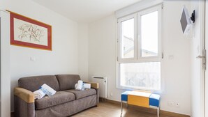 Un salon avec un canapé gris, une petite table surmontée d’un coussin bleu, un radiateur et une fenêtre avec des stores.