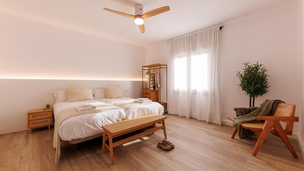 Ein modernes Schlafzimmer mit einem großen Bett, einer Holzbank, einem kleinen Tisch, einer Pflanze und einem Deckenventilator.