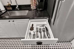 A kitchen with a black countertop, a stainless steel refrigerator, and a drawer with various utensils.