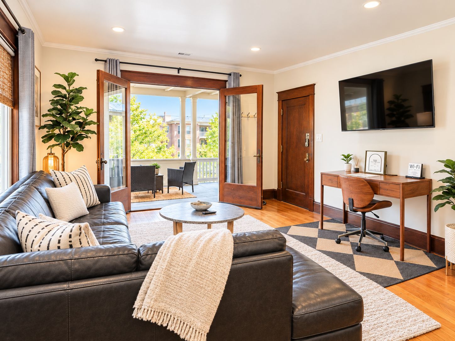 A modern living room with a leather sofa, a coffee table, a desk with a chair, a TV mounted on the wall, and a balcony with a view of buildings and trees.