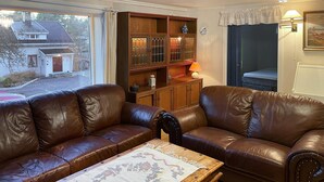 A living room with two brown leather sofas, a wooden coffee table with a floral table runner, a wooden cabinet with glass doors, and a window with a view of a house outside. - 2 bedroom lovely home in Færvik (Færvik)