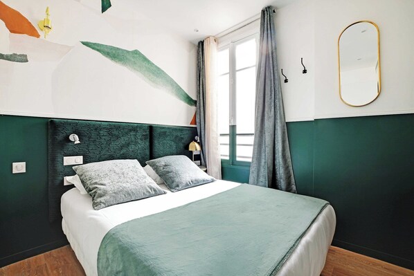 Ein ordentlich bezogenes Bett mit einem grünen Überwurf und Kissen, ein Kopfteil mit strukturierter Optik und ein großes Fenster mit Vorhängen.